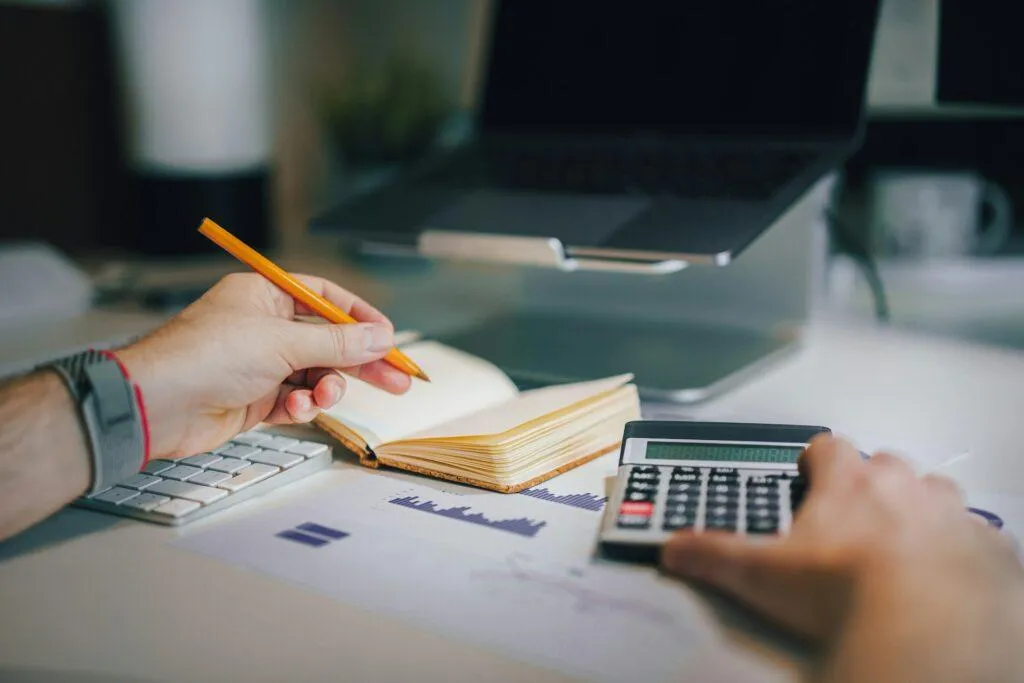 A man writes in a notebook while holding a calculator and viewing charts.